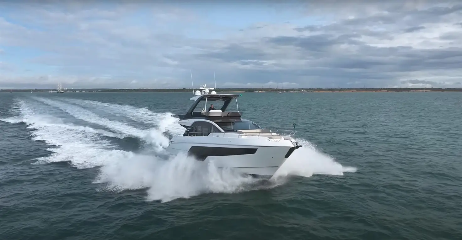 A luxury motor yacht speeds through blue waters, leaving a trail of white foam; clouds hover in a bright, partly cloudy sky.